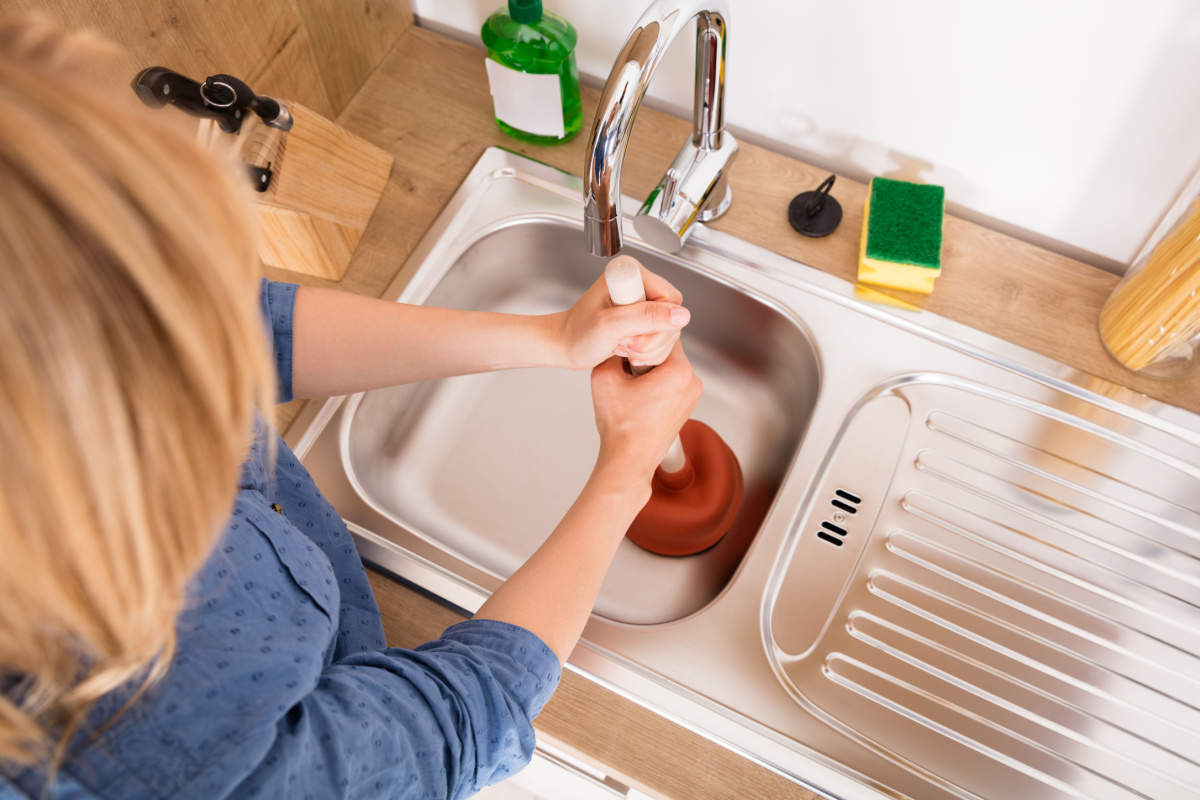 Woman fixing a clogged drain in her kitchen sink with a plunger